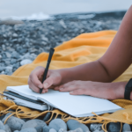 Person writing in a notebook on a rocky beach with a yellow blanket, planning a travel itinerary