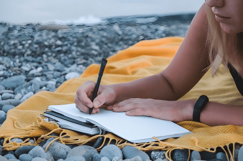 Person writing in a notebook on a rocky beach with a yellow blanket, planning a travel itinerary